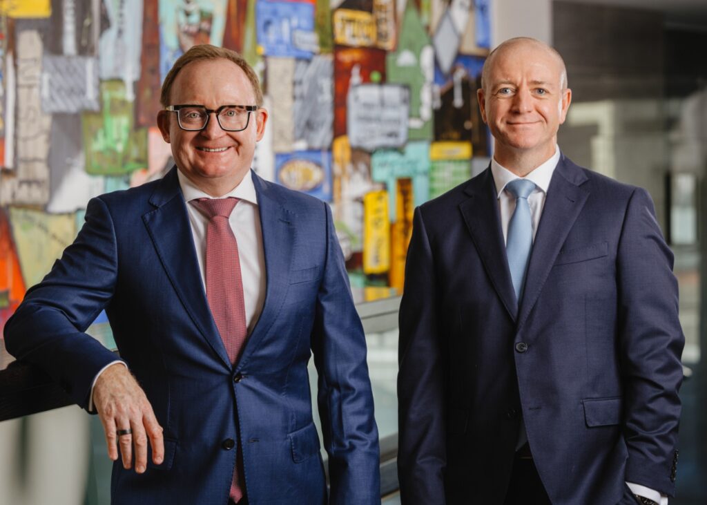 Two businessmen in navy suits stand side by side in a bright lobby with a colorful collage wall behind them.