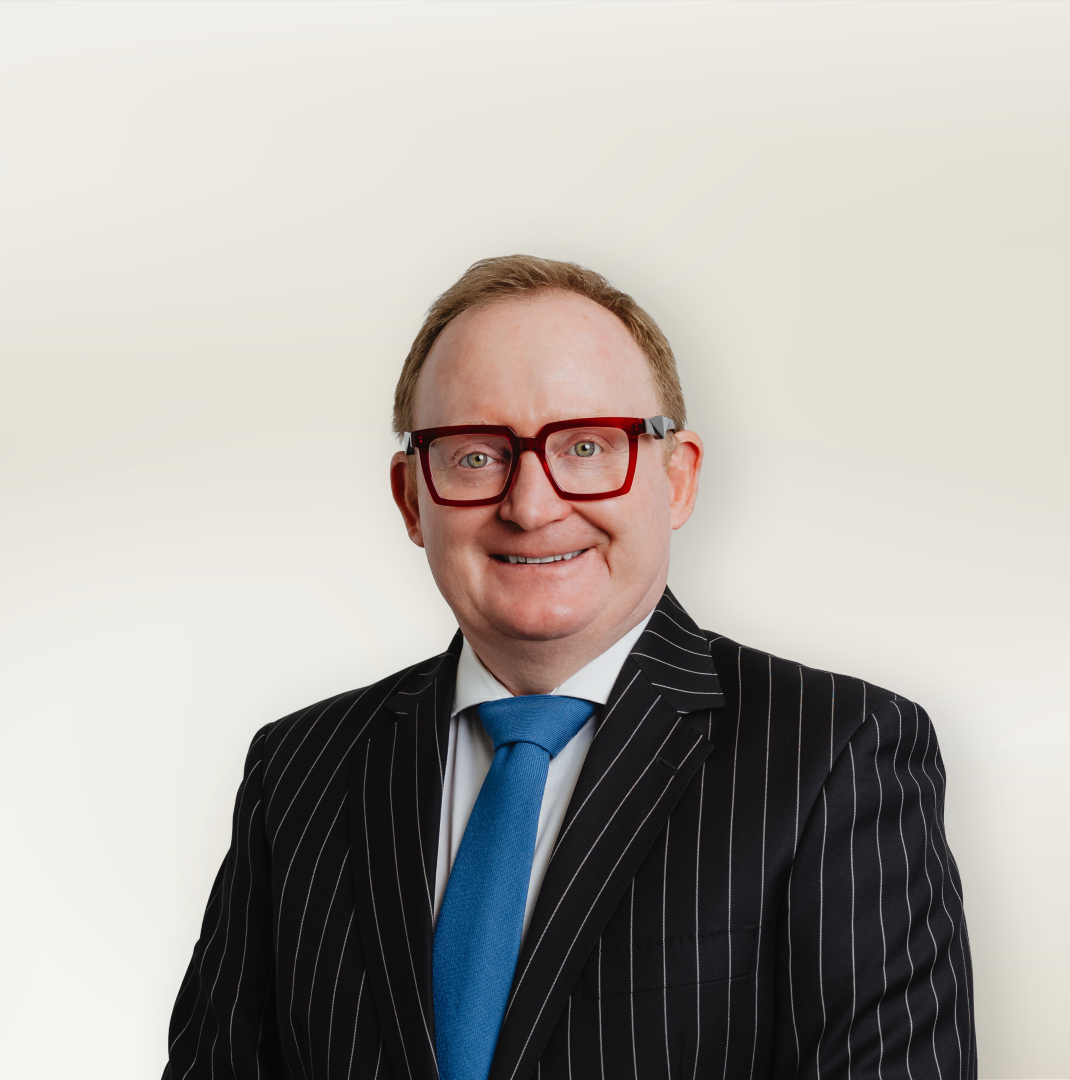 Smiling man in a dark pinstripe suit with a blue tie and red-framed glasses, posed against a light neutral background.