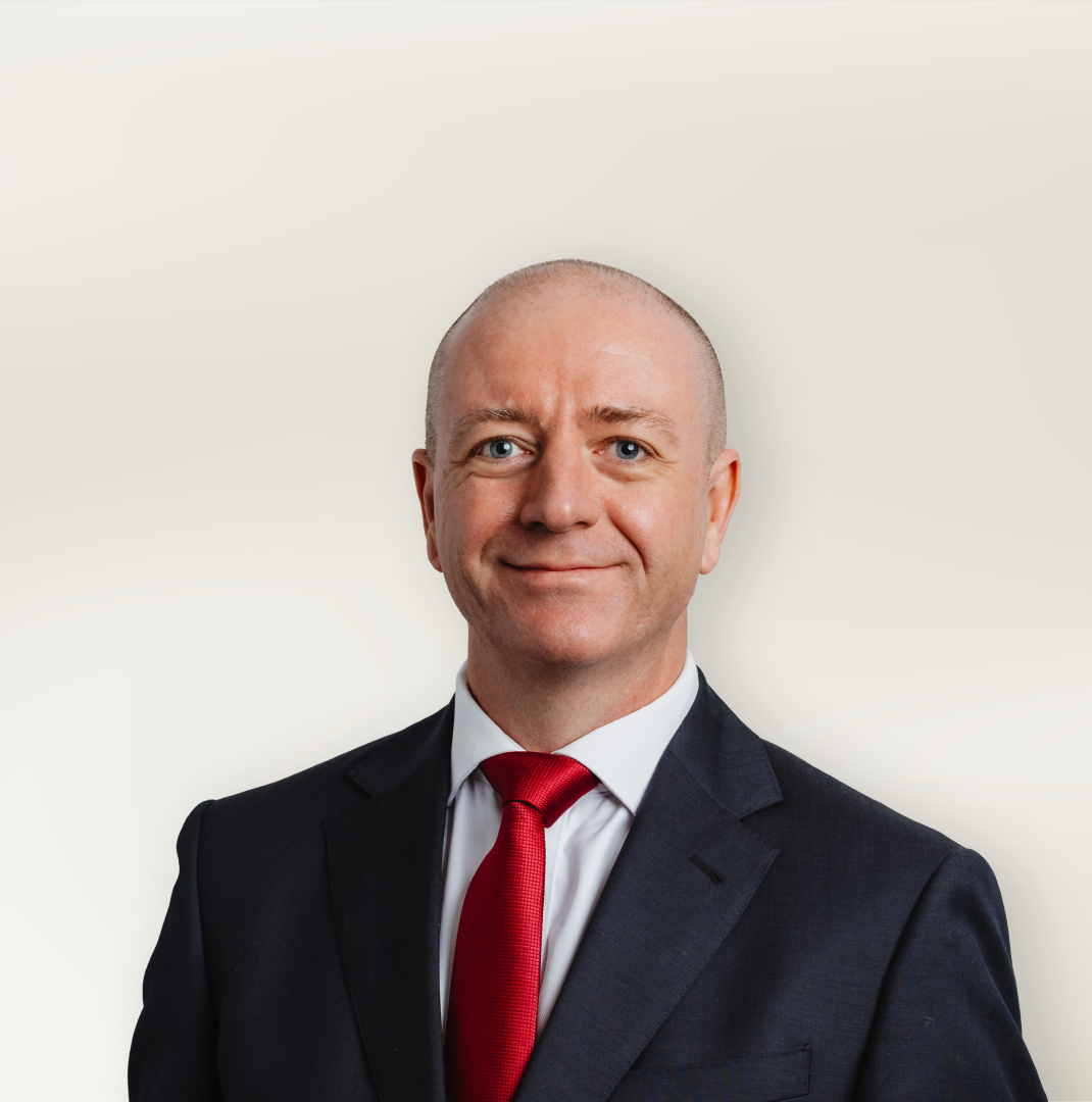 Professional headshot of a smiling, bald man in a dark suit and red tie against a light beige background on a studio backdrop.